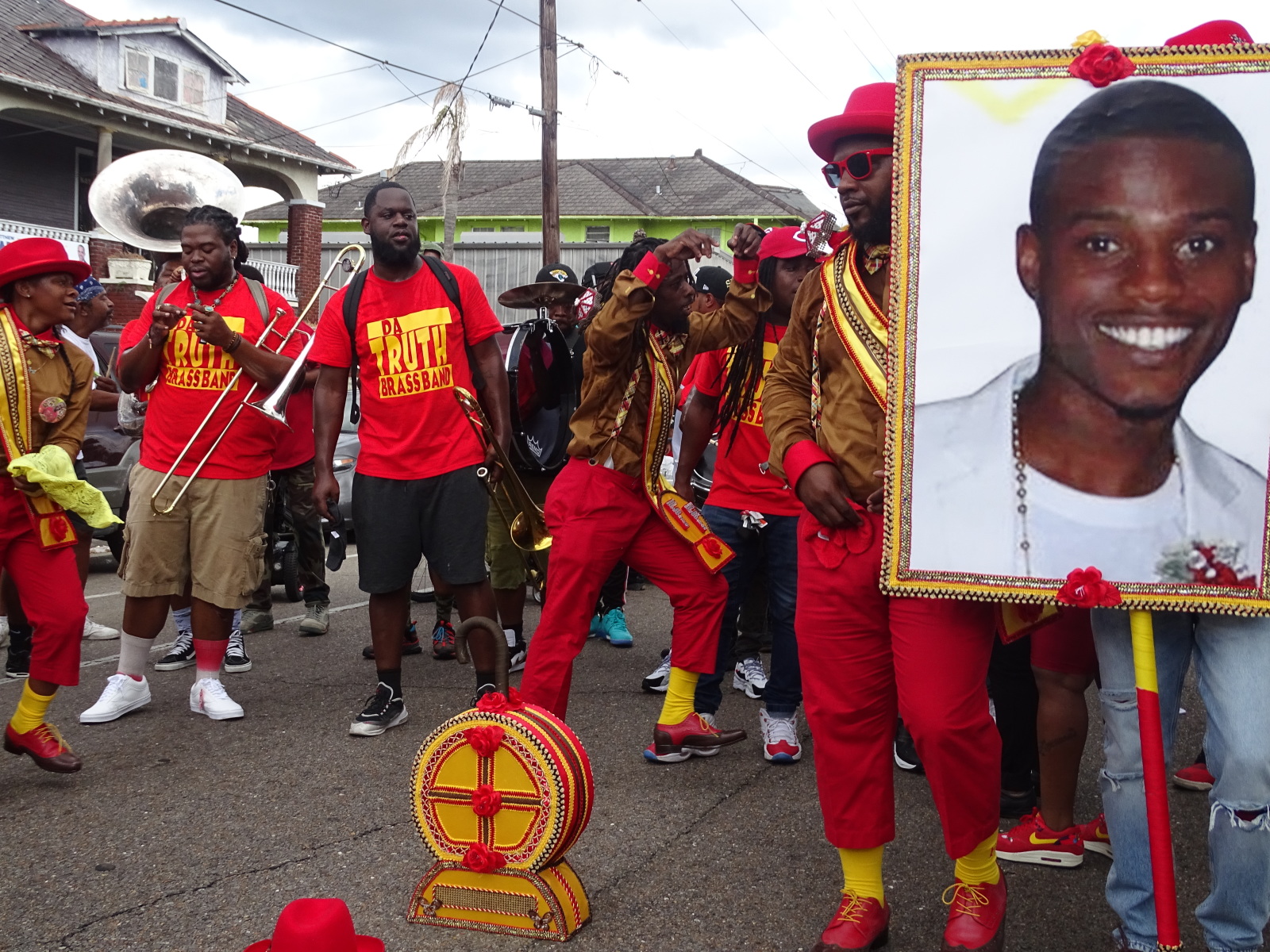 Derrick Santa Cruz memorializes Kharen Chapman with a large portrait photo during a Footwerk Family second line parade. A brass band can be seen in the background.
