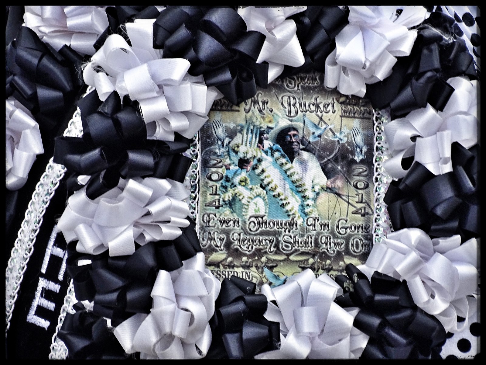 A memorial for Alfred “Bucket” Carter framed by black and white ribbon bows, featuring an image of a Carter labeled "Mr. Bucket" with the year "2014" and the phrase "Even Though I’m Gone, My Legacy Shall Live On." The design includes intricate details like musical instruments and decorative elements.