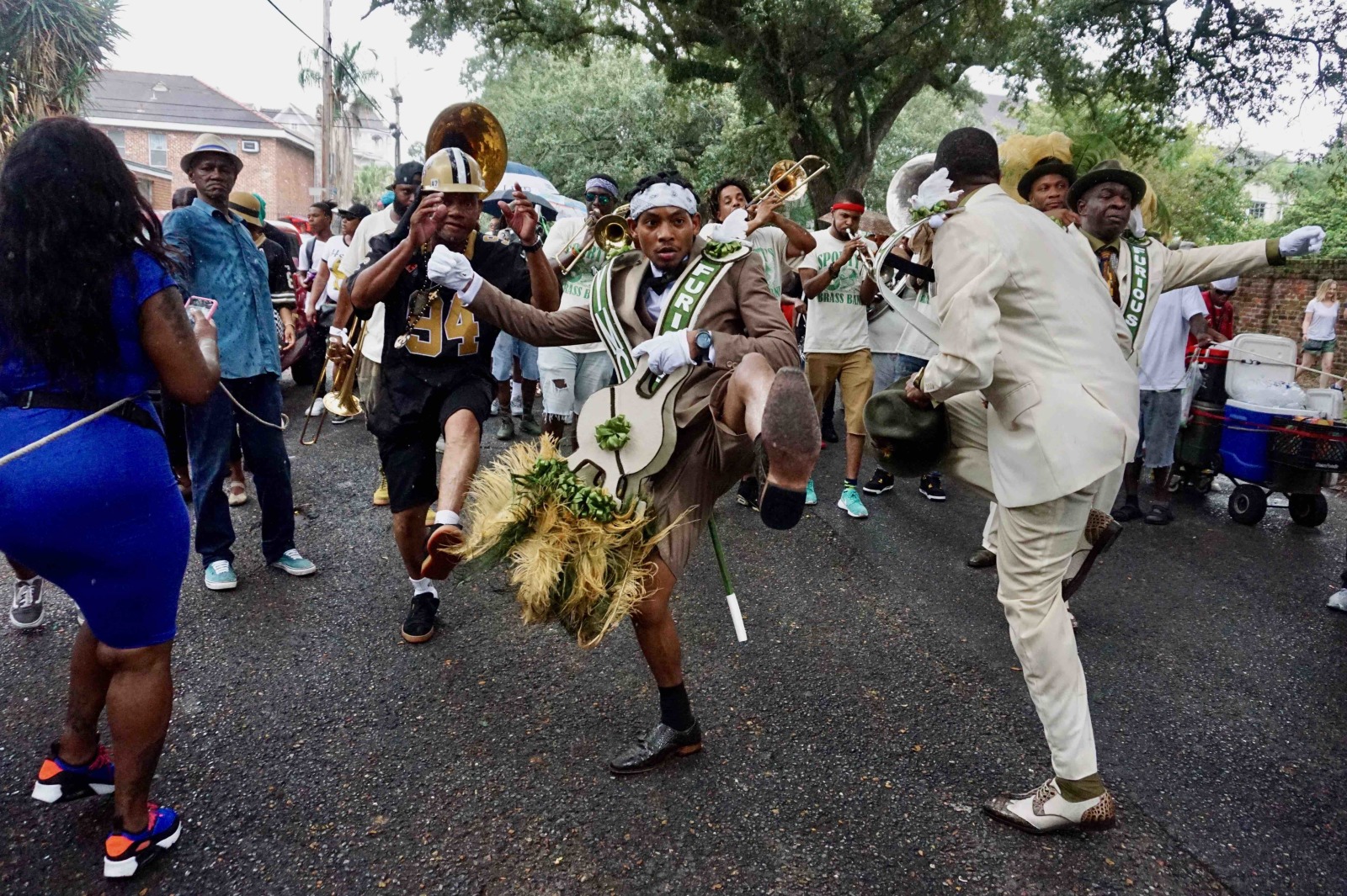 Waldorf Gipson IV is joined by several others in a second line parade, captured while kicking high in the air. They are wearing sashes and carrying decorated props, as a brass band plays behind them on a tree-lined street. A crowd of onlookers watches and captures the moment on their phones.
