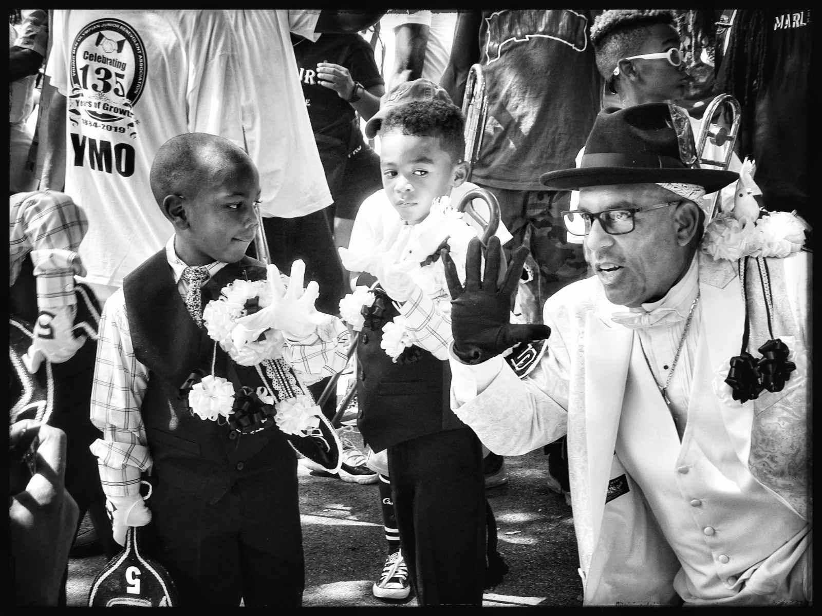 Two children, Eric Gordon Jr., Chance Gipson and Waldorf “Gip” Gipson, stand in the street at YMO Jr.'s 135th anniversary second line parade dressed in matching suits, as an older man gestures towards them.
