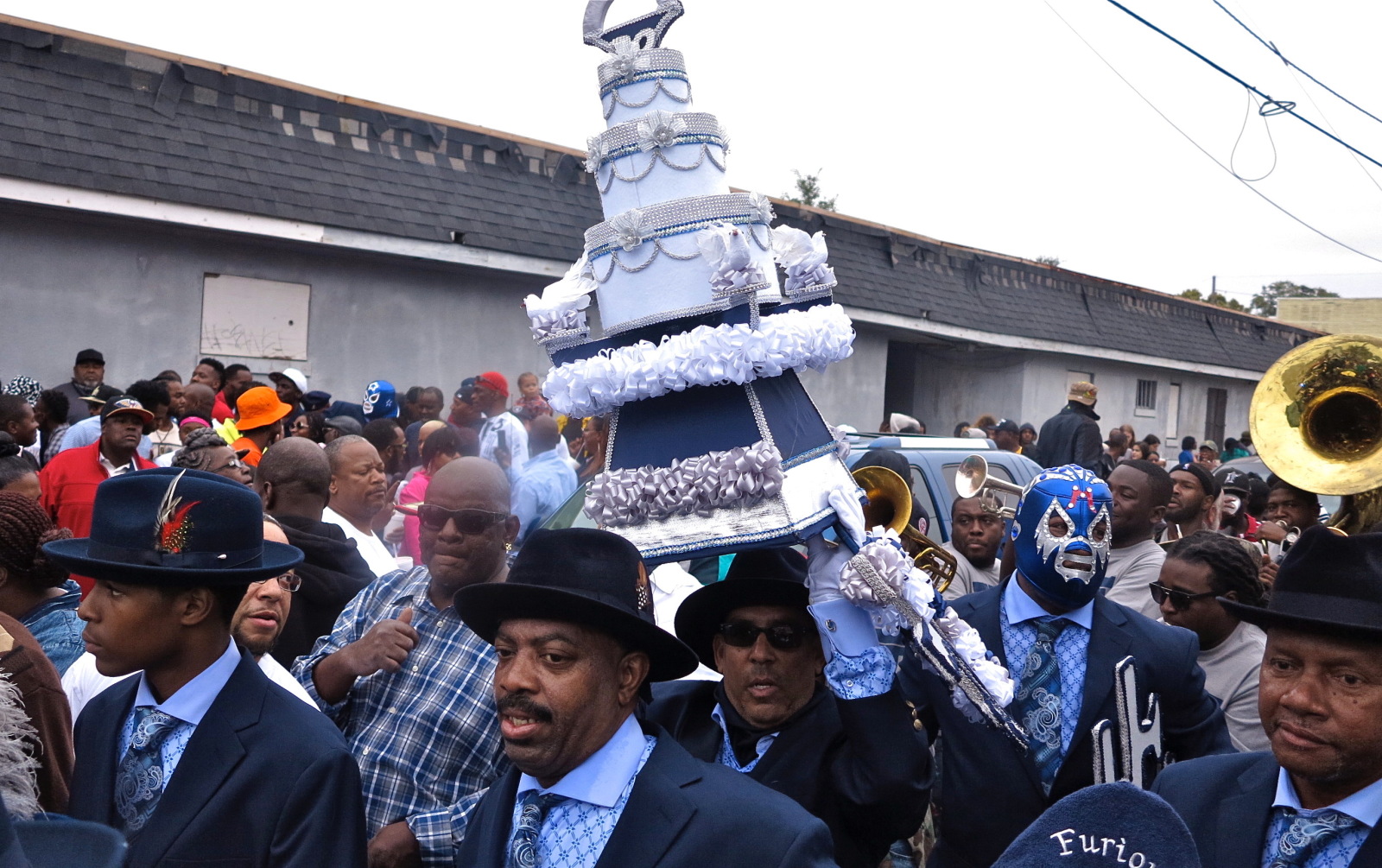 Furious Five founders Doo Right McCloud, Waldorf “Gip” Gipson, and Daniel Mason (in mask) march in a parade wearing coordinated blue suits and ties. Gipson carries an elaborate, cake-like decoration through a crowded street, accompanied by brass musicians and onlookers.