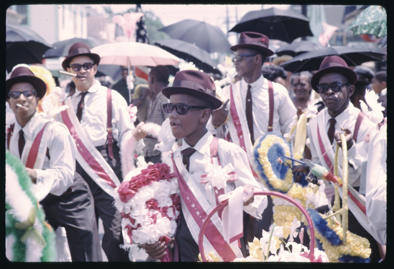Parade participants dressed in coordinated white shirts, red suspenders, and brown hats march through a lively crowd. They carry colorful wreaths and decorations, with umbrellas dotting the background.
