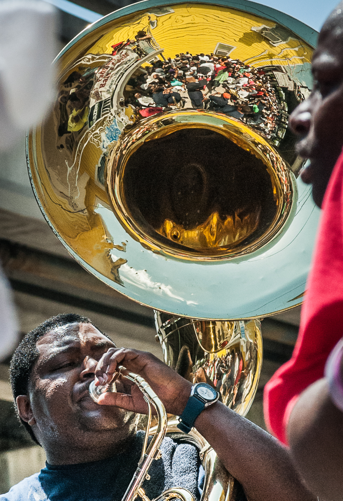 Bennie Pete, leader of the Hot 8 Brass Band, plays sousaphone at a second line parade, his face focused as he blows into the brass instrument. The shiny bell reflects a lively crowd gathered in front of a yellow building.