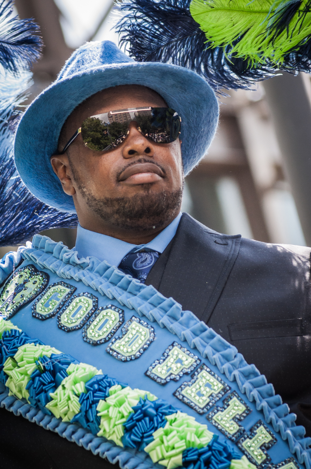 Raynold Fenelon, member of the Good Fellas, dressed in a blue suit, matching hat, and sunglasses confidently participates in a parade. His sash, decorated with ruffles and beads, reads "GOOD FELLAS," and vibrant feathers extend from his outfit.