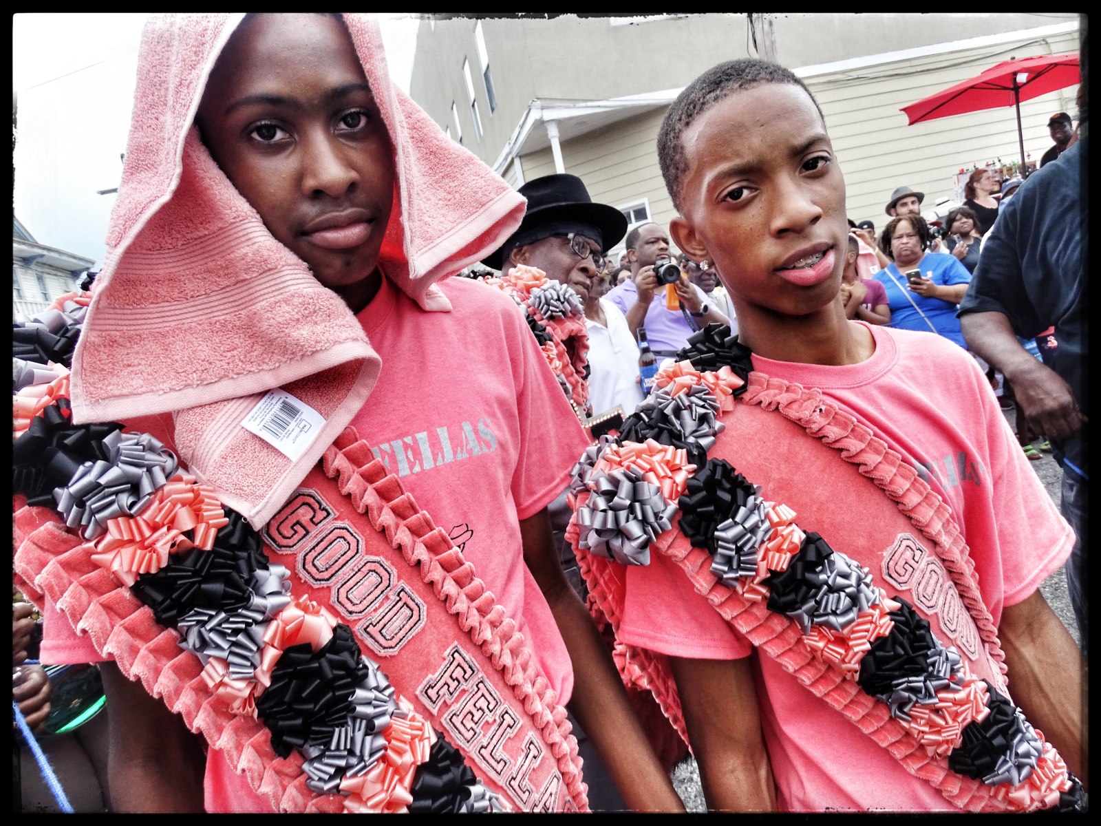 Two young men, Raynold Fenelon, Jr. and Hassan Goffner, wear matching pink shirts and elaborately decorated sashes reading "GOOD FELLAS" and stand amidst a lively crowd. One drapes a pink towel over his head.