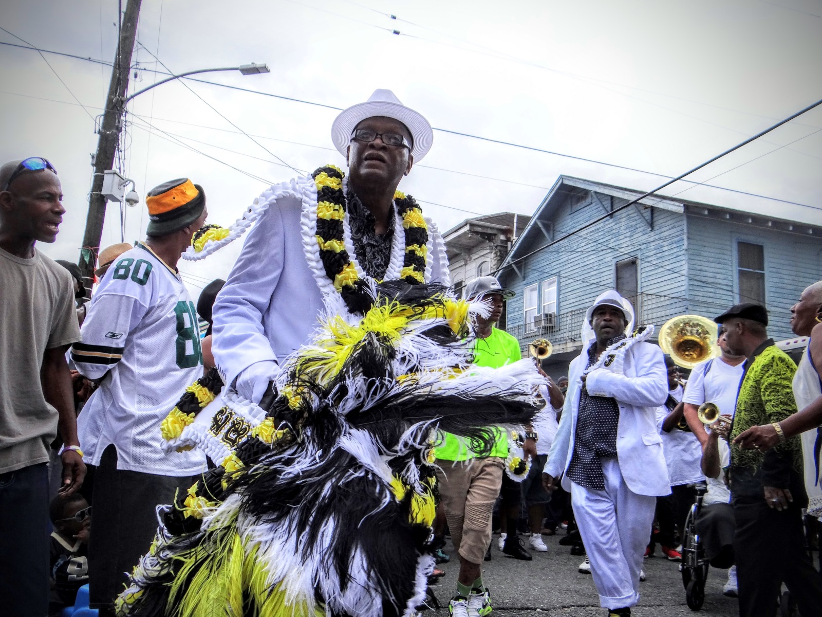Albert Clark, dressed in a white suit, hat, and a sash adorned with black and yellow feathers, walks in a lively street parade. Behind him, a brass band and a crowd of spectators follow.