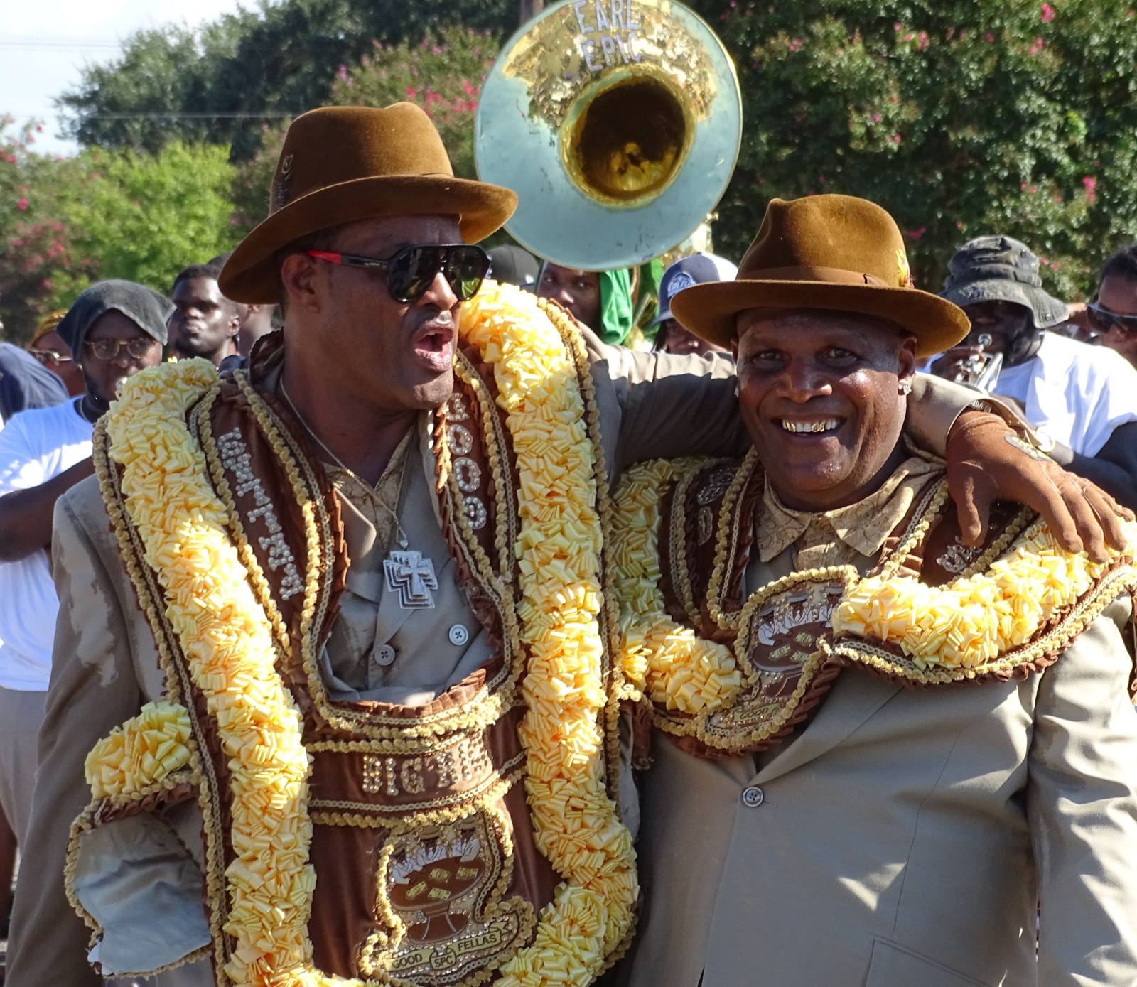 Richard Brown and Steven Hill wear streamers, matching brown suits, wide-brimmed hats, and elaborately decorated yellow and brown sashes celebrate together at a parade. One has his arm around the other as they smile and cheer, with a brass band playing in the background.