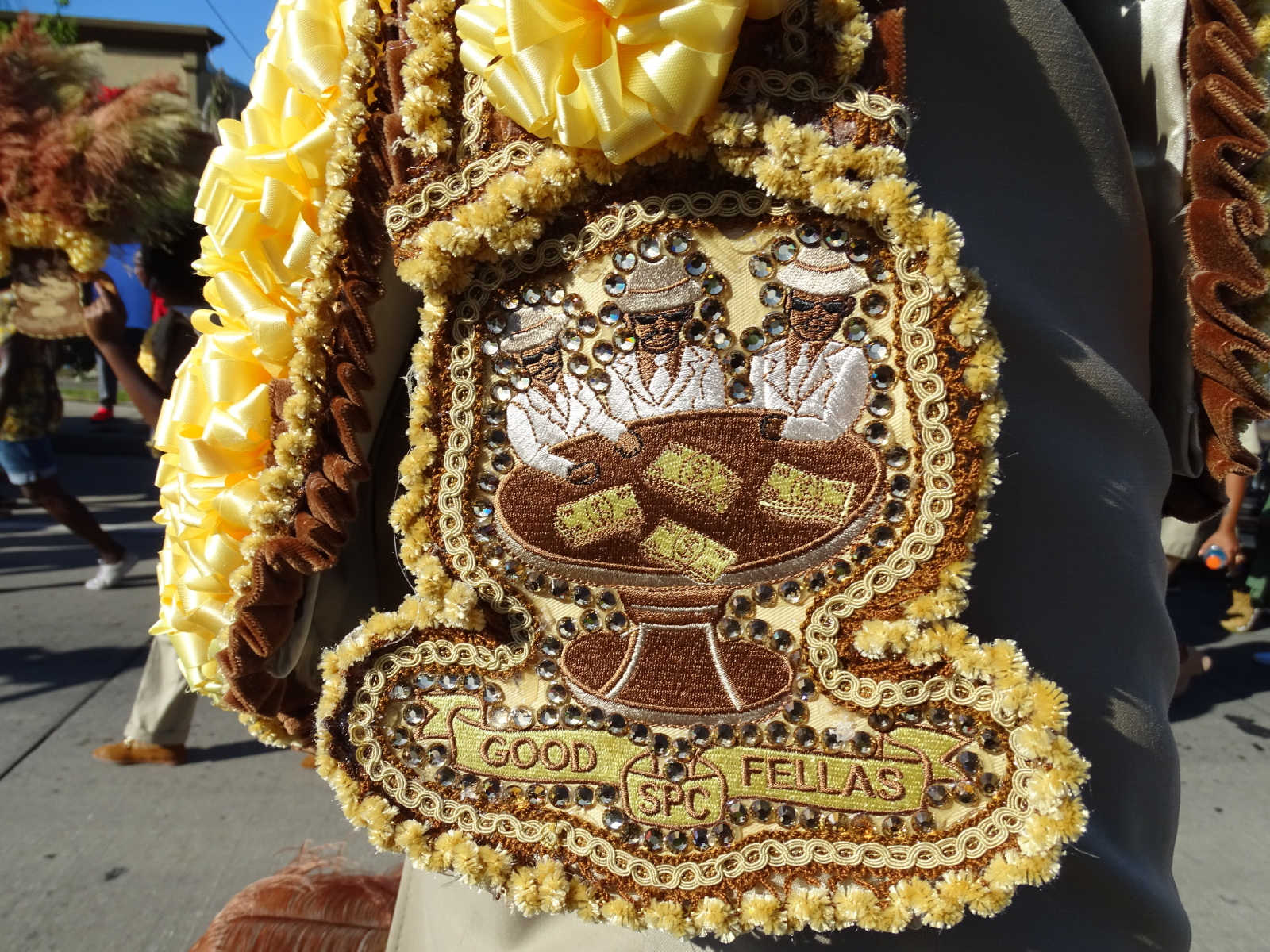 A close-up of an ornate streamer featuring intricate beadwork, yellow ribbon accents, and an embroidered design. The patch depicts two men in white suits and sunglasses sitting at a table with stacks of money, with the words "GOOD FELLAS SPC" stitched below.