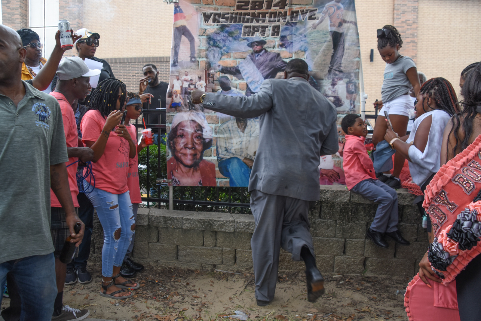 Raynold Fenelon wears a gray suit and dances energetically in front of a large memorial banner featuring images of his mother. A crowd of onlookers, some in matching pink shirts, watches and celebrates around him.
