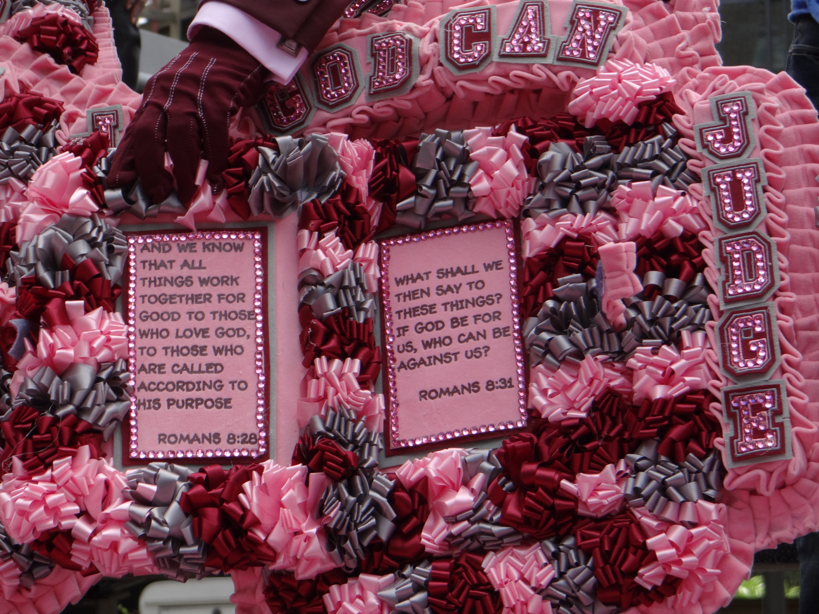 A close up of Good Fellas tenth anniversary regalia, made by Kevin Dunn. A decorative pink and red wreath adorned with ruffled fabric and bows features two framed Bible verses from Romans 8:28 and 8:31. The top of the wreath displays the phrase "GOD CAN JUDGE" in bold, glittery letters.
