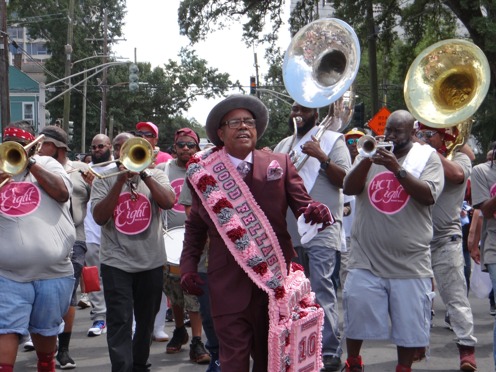 Kevin Dunn wears a burgundy suit, hat, and gloves leads a lively parade, wearing a pink and red sash that reads "GOOD FELLAS" and carrying a matching decorative piece. Behind him, the Hot 8 brass band plays.