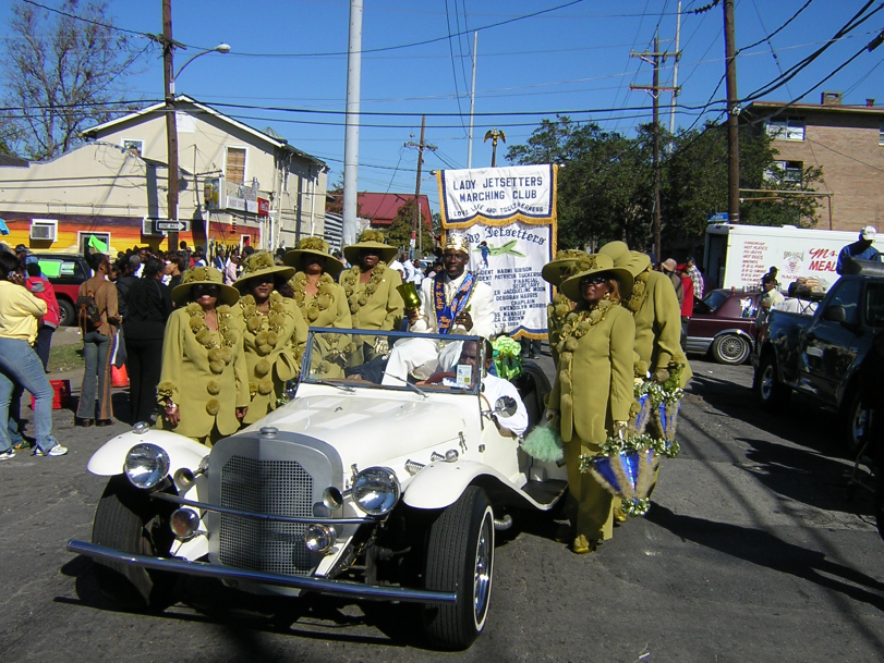 Members of the Ladies Jetsetters wearing matching yellow-green suits and hats stand around a vintage white convertible during a street parade. Greg Stafford, in a decorated sash, sits in the car with a banner reading "Lady Jetsetters Marching Club" in the background.