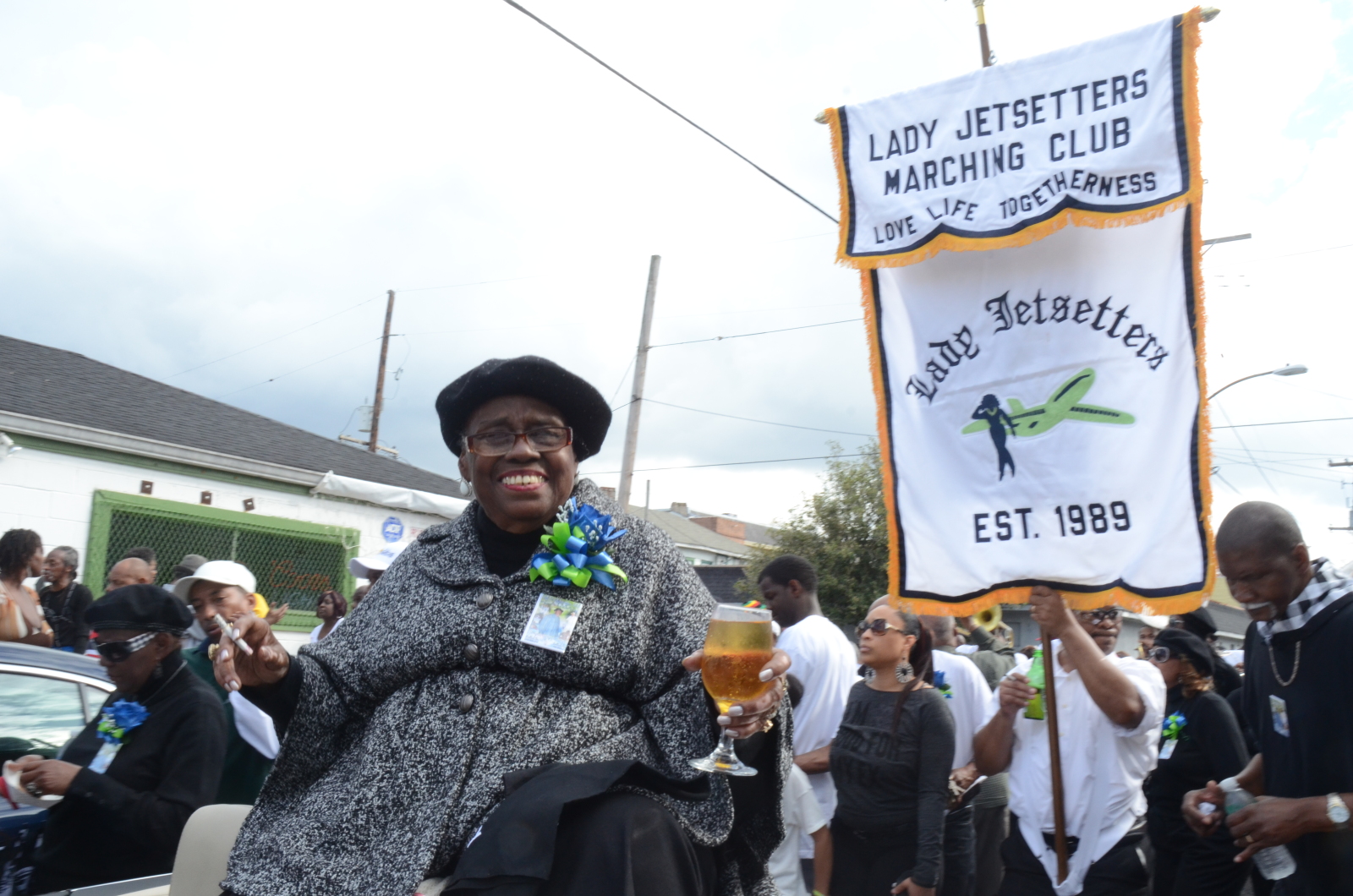 Naomi Gibson, wearing a black hat and gray coat, sits in a parade car, holding a drink and smiling. A banner reading "Lady Jetsetters Marching Club, Est. 1989" hangs in the background, surrounded by people in festive attire.