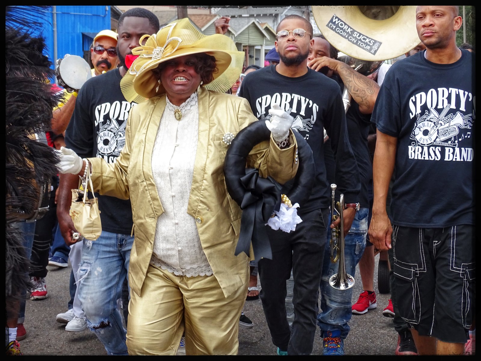 Creola “Jackie” Moon honors Naomi Gibson at the Lady Jetsetters 30th anniversary parade. she's wearing a gold suit, pants, and matching large hat. A brass band follows behind her.
