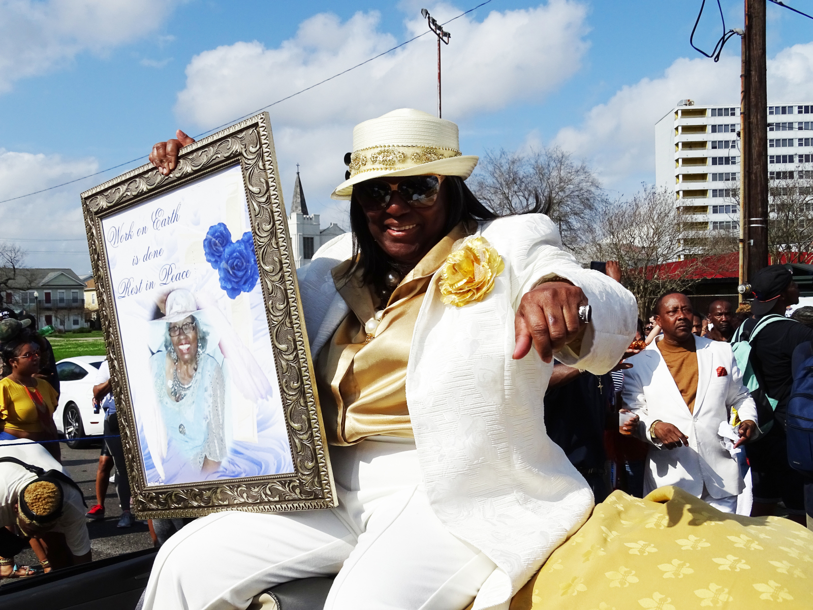 Shelia Gibson honors her mother, Naomi Gibson, at the Lady Jetsetters 30th anniversary parade. She is wearing a white suit with gold shirt and hat, and is riding in an open-top convertible.