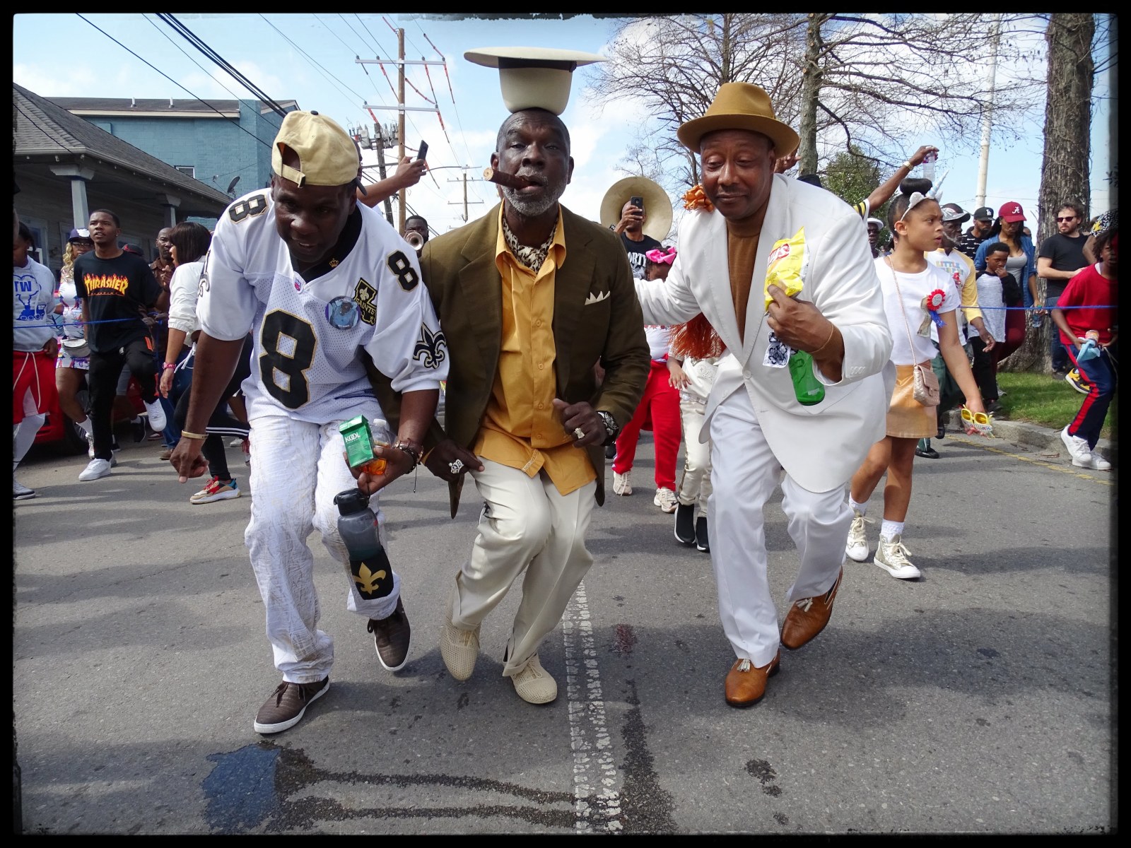 Three men—Roque “Rock” Gibson, Gregg Stafford, and Fredrico—dance side by side at the Lady Jetsetters 30th anniversary parade. A brass band and parade attendees can be seen behind them.