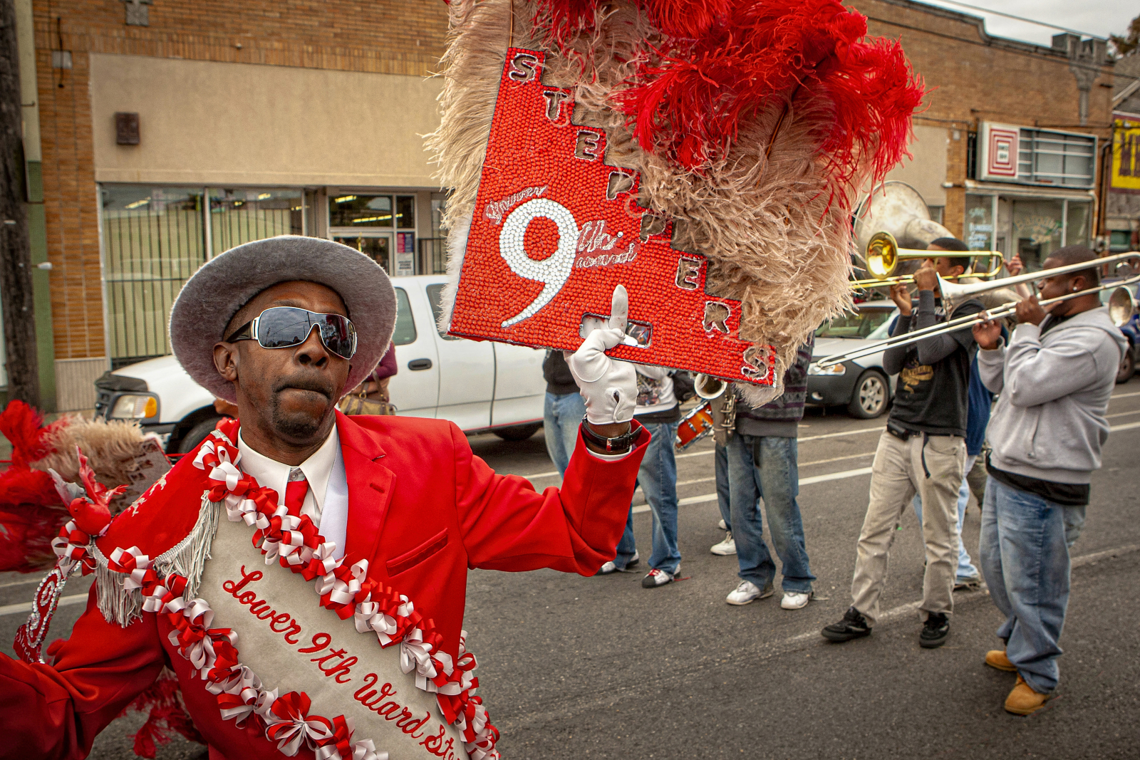 A man in a bold red suit, white gloves, and a silver hat dances energetically, holding a red and beige feathered fan with "Lower 9th Ward Steppers" written on his sash, as a brass band plays behind him.