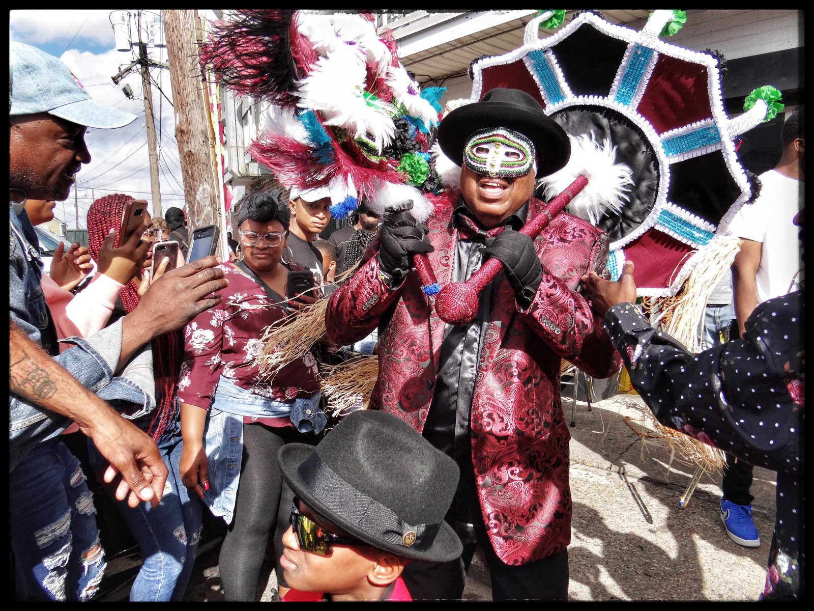 A man in a red and black paisley suit, black hat, and decorative mask dances energetically in a street parade, holding an ornate feathered umbrella, as a crowd watches and captures the moment on their phones.