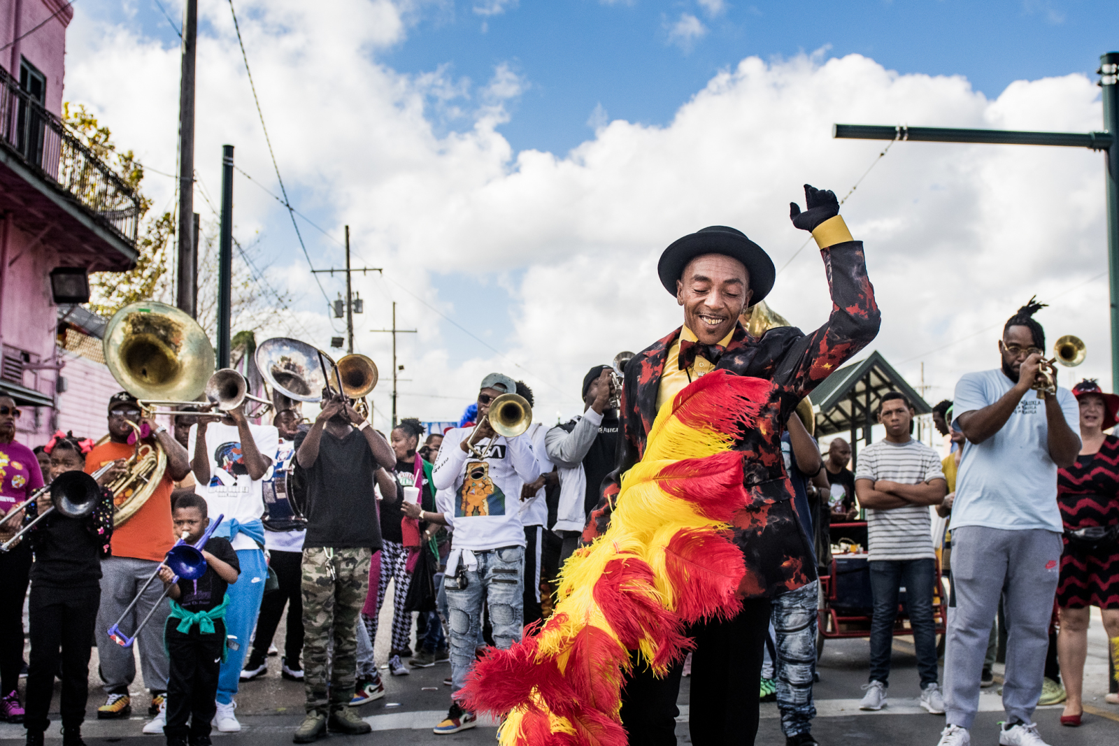 A joyful dancer in a black and red suit with a bright red and yellow feathered sash moves energetically through the street, leading a brass band performing in a lively parade.