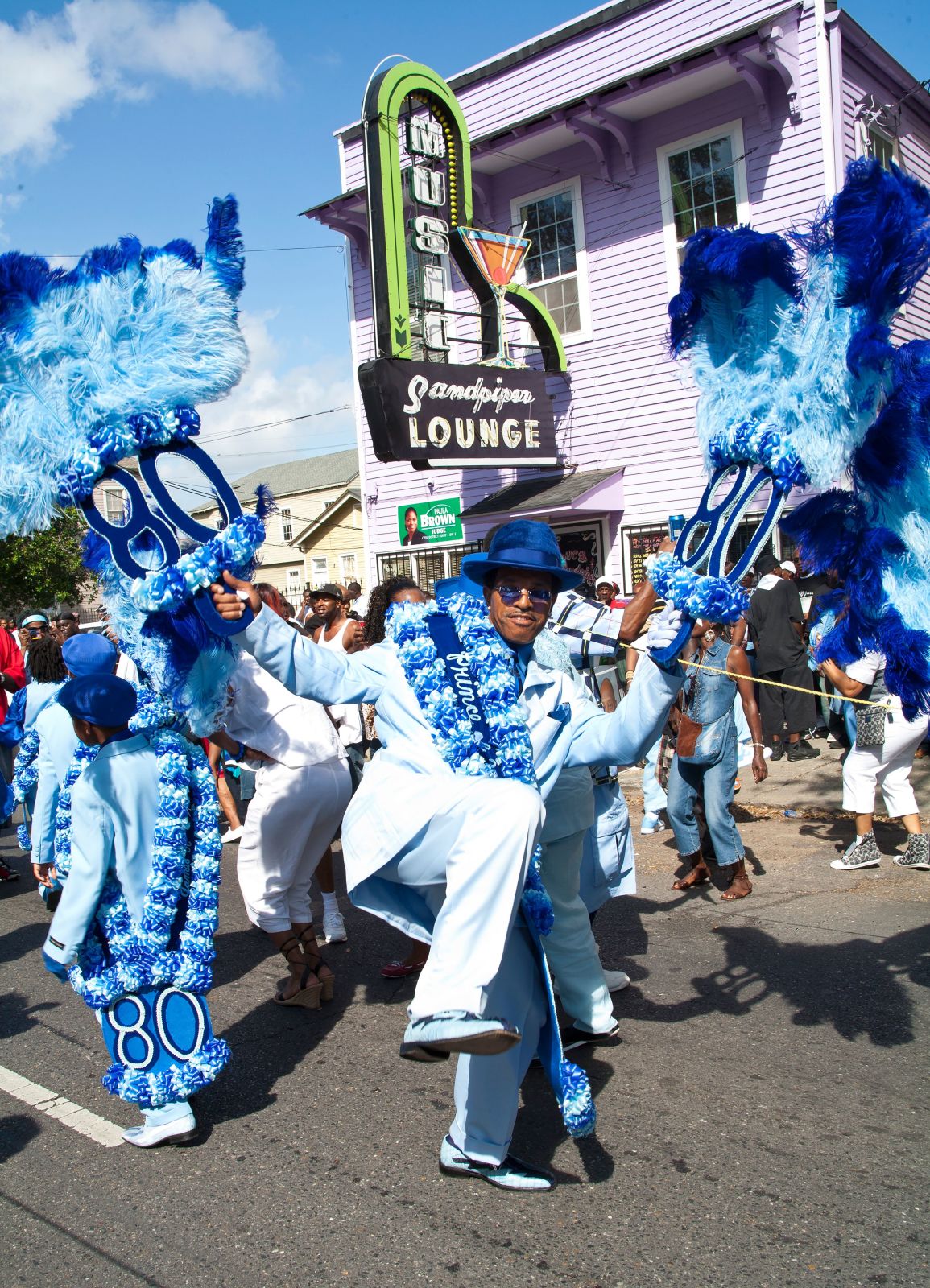 Alvin “Quiet” Epps, wearing a crisp white suit with blue sash and hat, dances with large feather parade props. The marquee sign for the Sandpiper Lounge is pictured in the background.