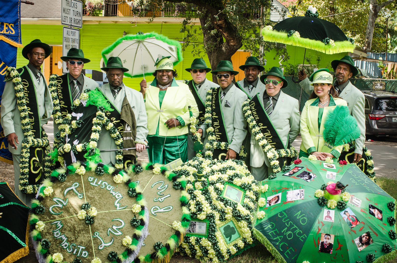 Members of Prince of Wales, dressed in bright green and black suits, pose in front of decorated parasols adorned with names of members and civil rights phrases including "NO JUSTICE, NO PEACE."