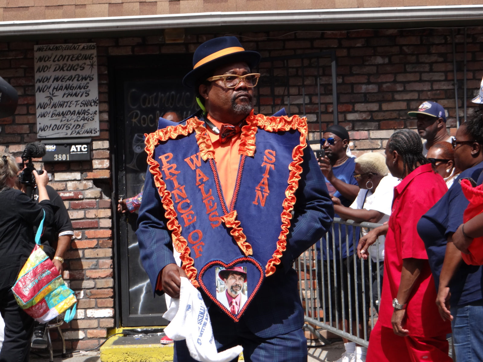 Stanley Taylor stands outside the Rock Bottom Lounge at a second line parade, wearing a purple suit, hat, and sash, with orange accents.
