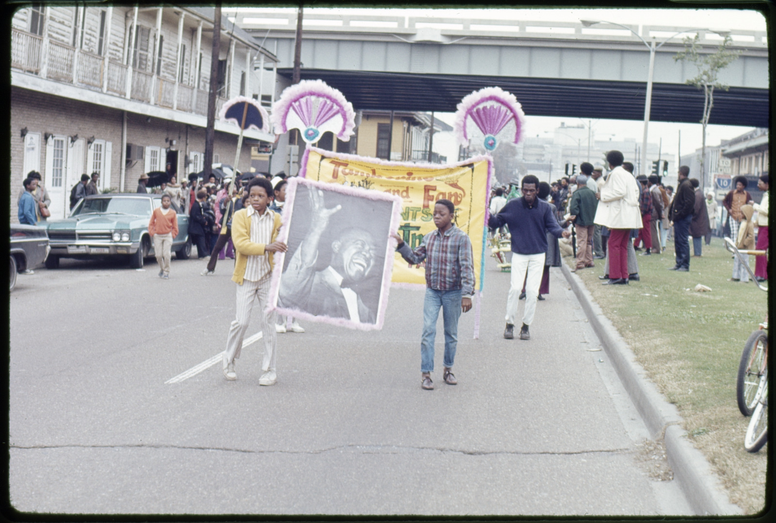 A vintage color photo from 1971 shows members of a Tamborine and Fan parade coming down the street. The Claiborne overpass can be seen in the background.