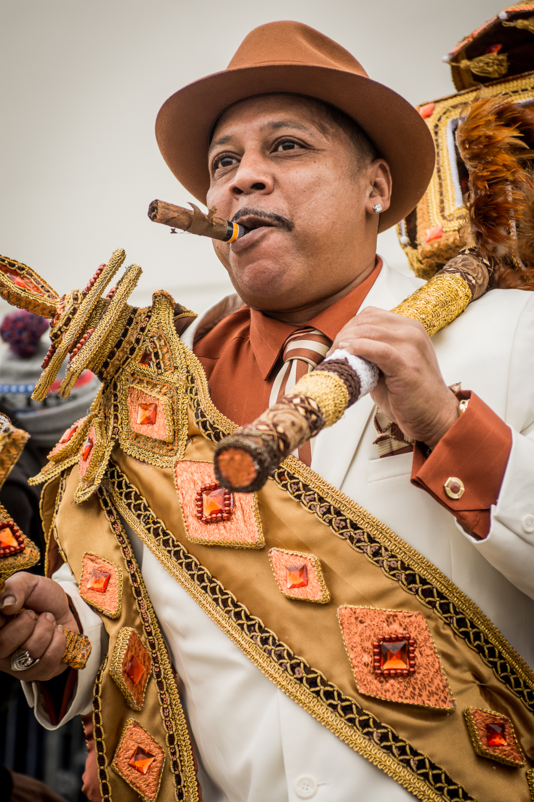 Tyrone "Pie" Stevenson of Spirit 2 Da Street. He wears an elegant white suit and brown hat, and smokes a cigar while leading a second-line parade. He wears a sash adorned with gold and orange embellishments, holding a matching decorated staff.