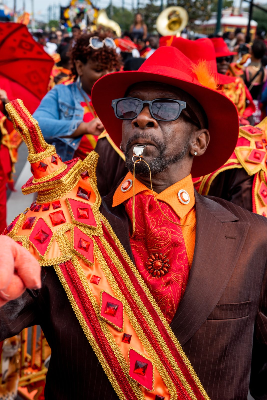 Larry Terrance with Spirit 2 Da Street. He's wearing a maroon jacket with orange undershirt and red and orange decorated sash and orange hat. He marches down the street with a whistle in his mouth.