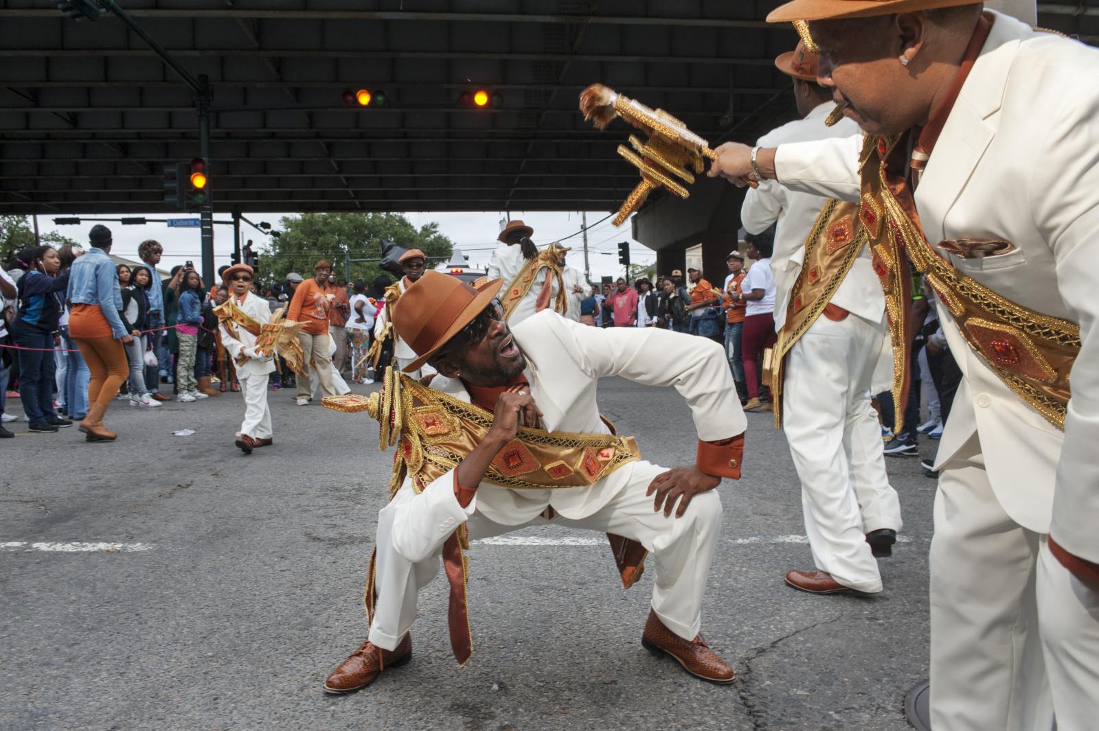 Larry Terrance and Tyron “Pie” Stevenson dance at a second line parade. They wear all-white suits with orange decorated sashes. The Claiborne overpass can be seen in the background.