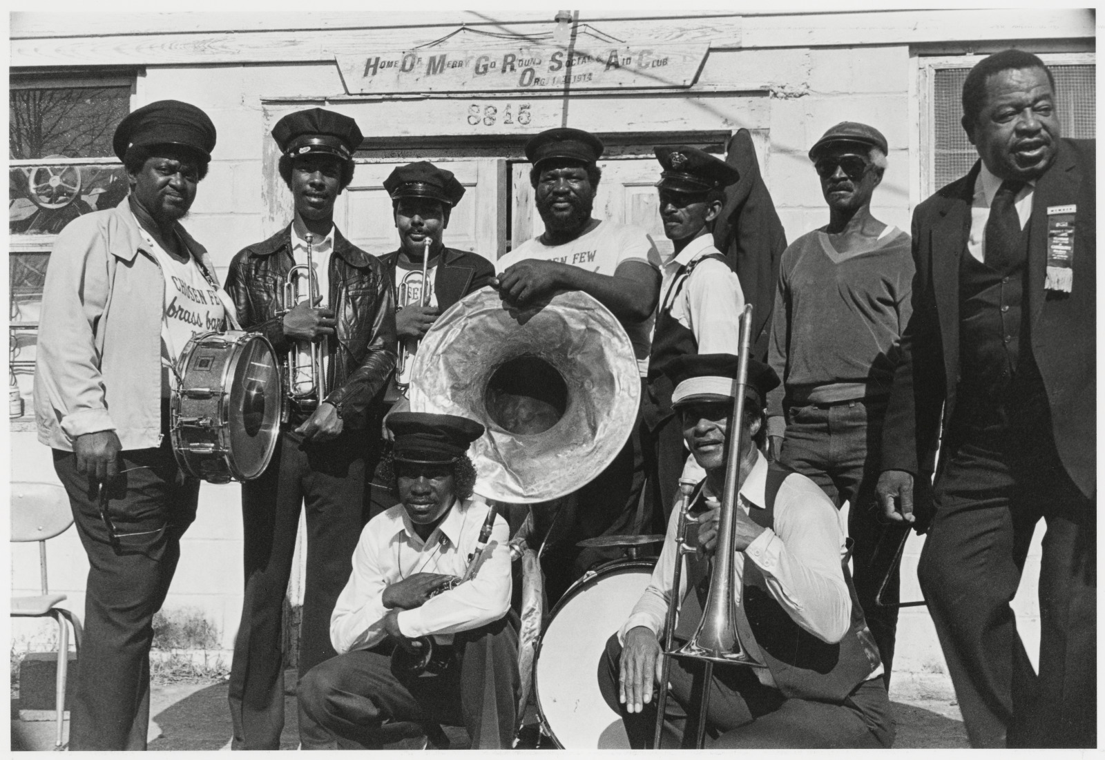 View of a group of band members posed for the photographer outside of a building located at 8815 Hickory Street. A wooden sign with the words Home of Merry Go Round Social Aid Club Organized 1914 can be seen above the double doors where the street number 8815 is also seen. The band shown is the Chosen Few Brass Band formed by Anthony "Tuba Fats" Lacen in the mid 1970s.