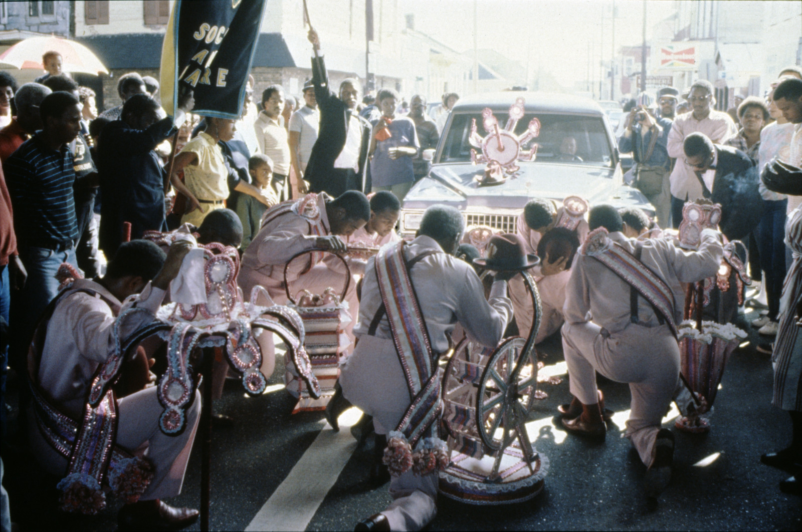 A vintage color photo shows members of Sudan, wearing matching white outfits while kneeling in the street, pay respects to the late Archie Chapman, whose basket can be seen on the front of the hearse.