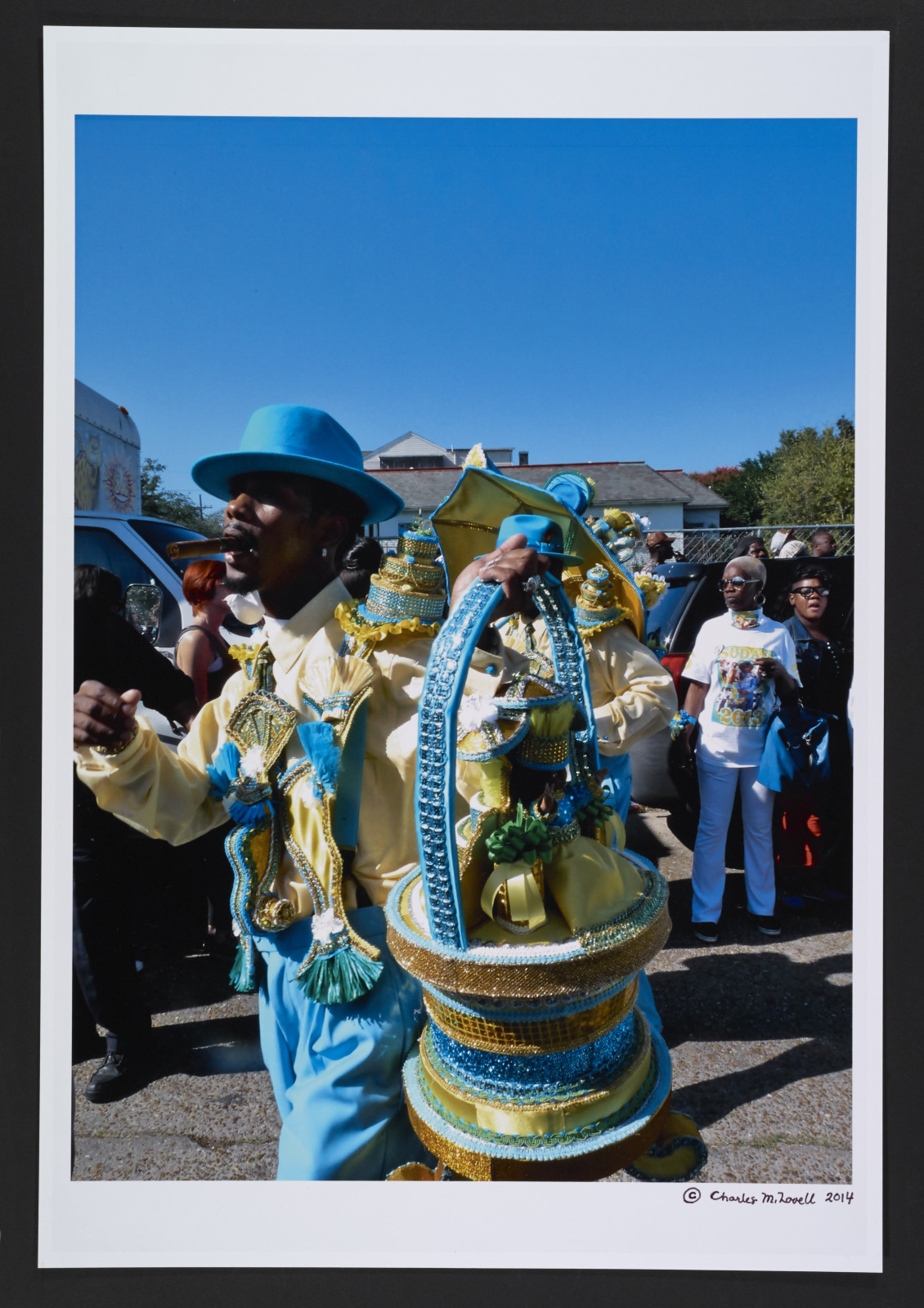 View a Sudan second line parade in the Treme neighborhood. The view features a club member wearing robin egg blue slacks and hat, a yellow shirt, and carrying a large decorative basket.
