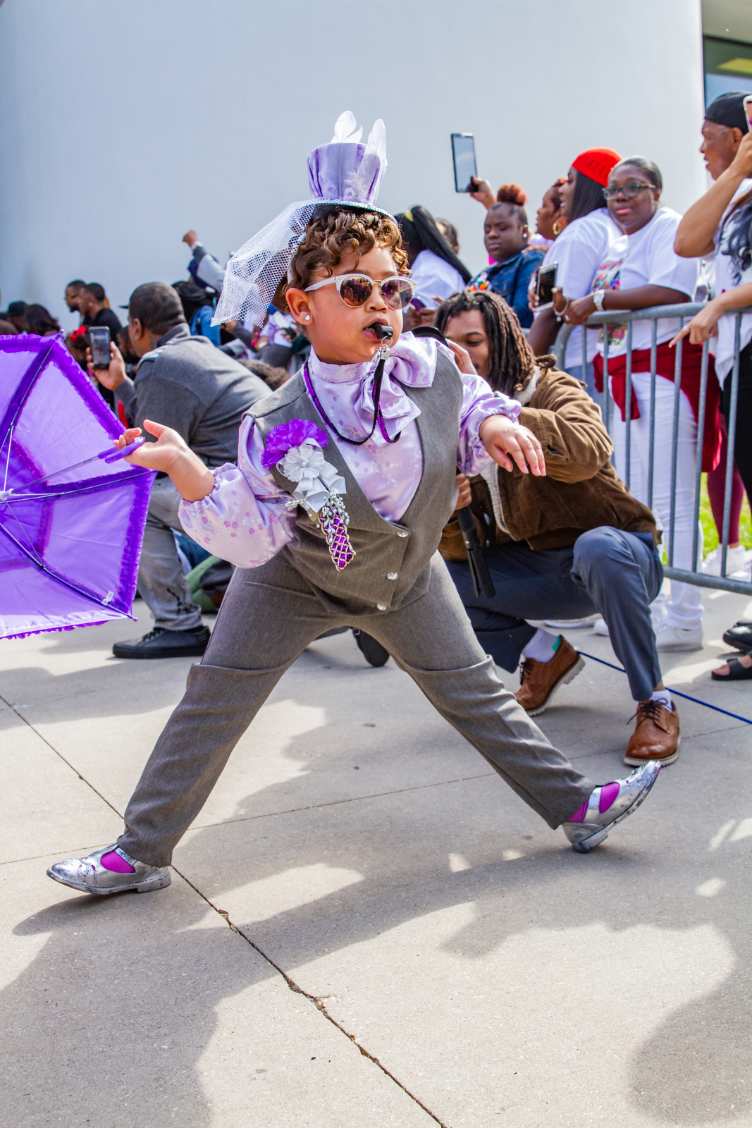 Sage Santa Cruz comes out of the door at a second line parade dressed in a suit and holding a purple parasol with a whistle in her mouth.