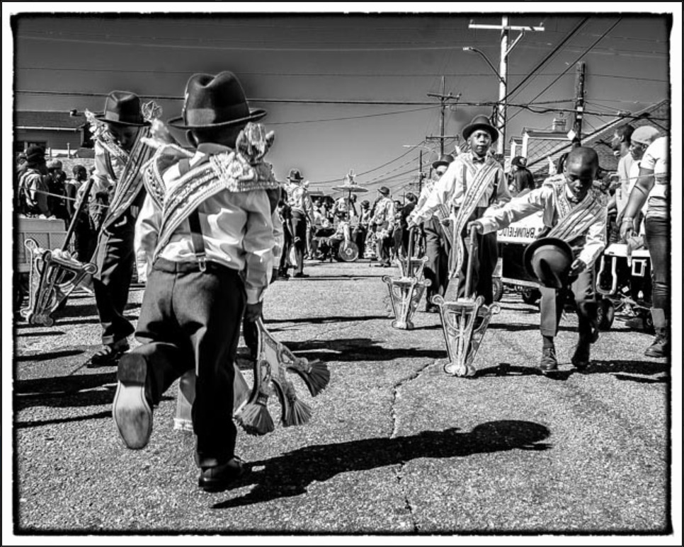 A black and white photo shows the children's division of Sudan at a second line parade, with its members dancing with parade props.