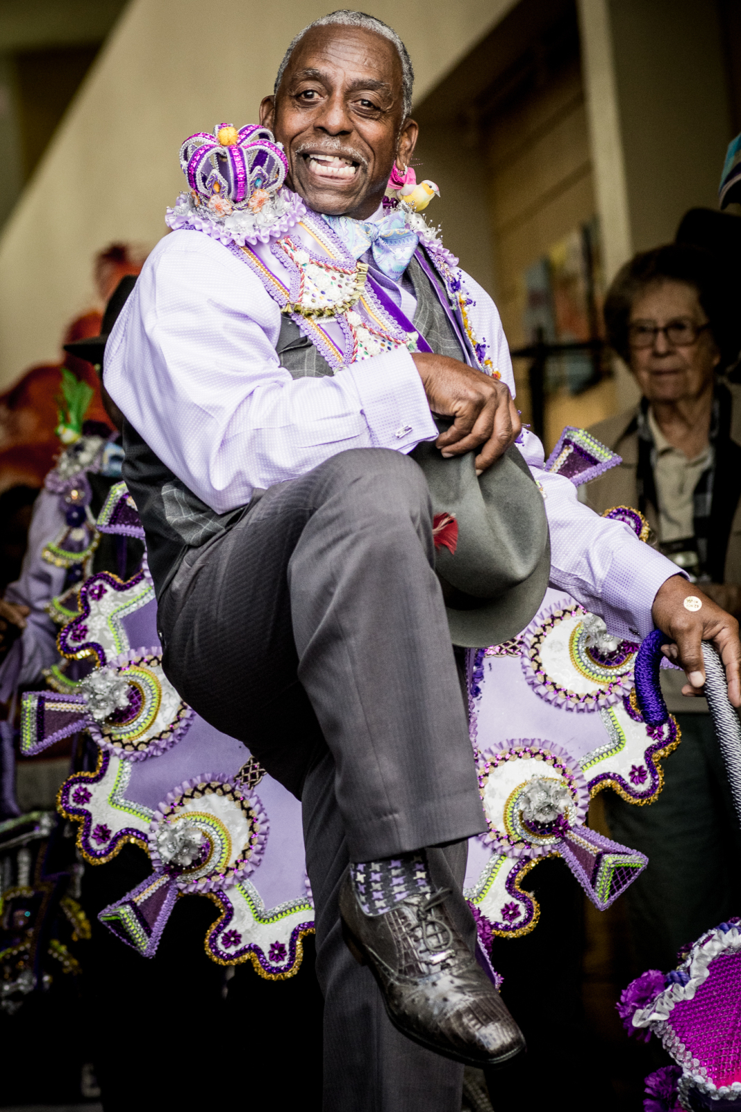 Bernard Robinson of the Sudan social aid and pleasure club, high steps down the street at a second line parade wearing a blue and grey suit, white shirt, wingtip shoes, and a bowler hat, He's wearing a decorated purple sash with matching parasol.