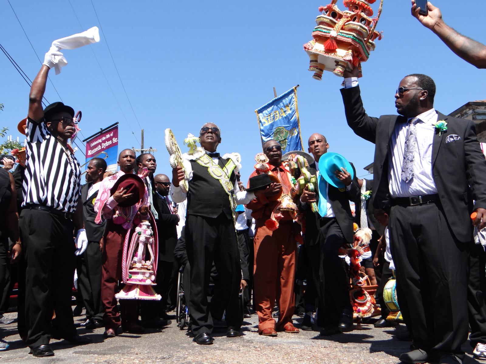 A view of the second line parade funeral for Kenneth Dykes in 2015. Members of Sudan can be seen wearing black suits, white shirts, and multicolored ties, carrying parade banners and props.