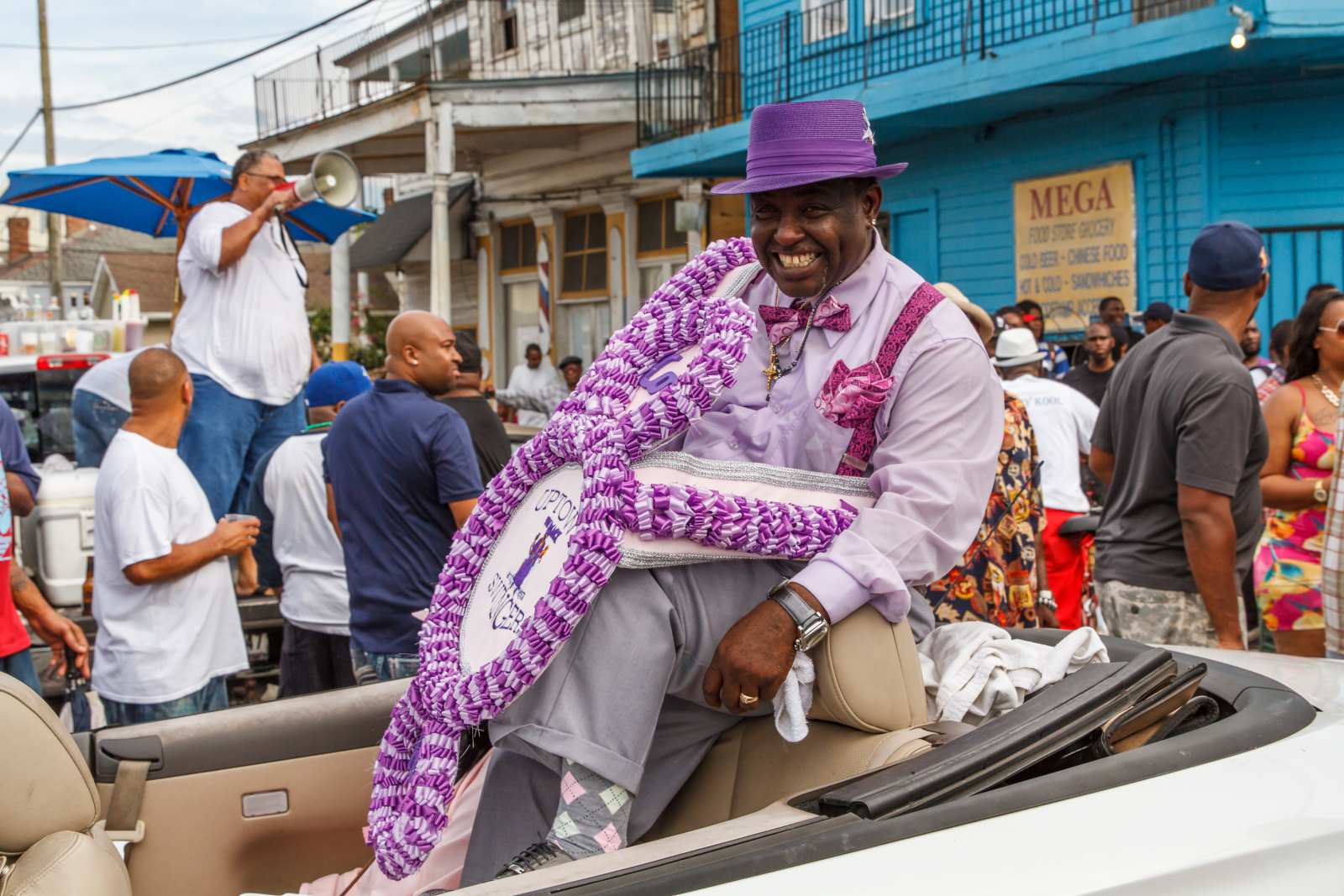 A joyful man dressed in a coordinated lavender and purple outfit, complete with a matching hat, bow tie, and suspenders, sits atop a convertible during a lively street parade, holding an elaborately decorated second-line accessory.