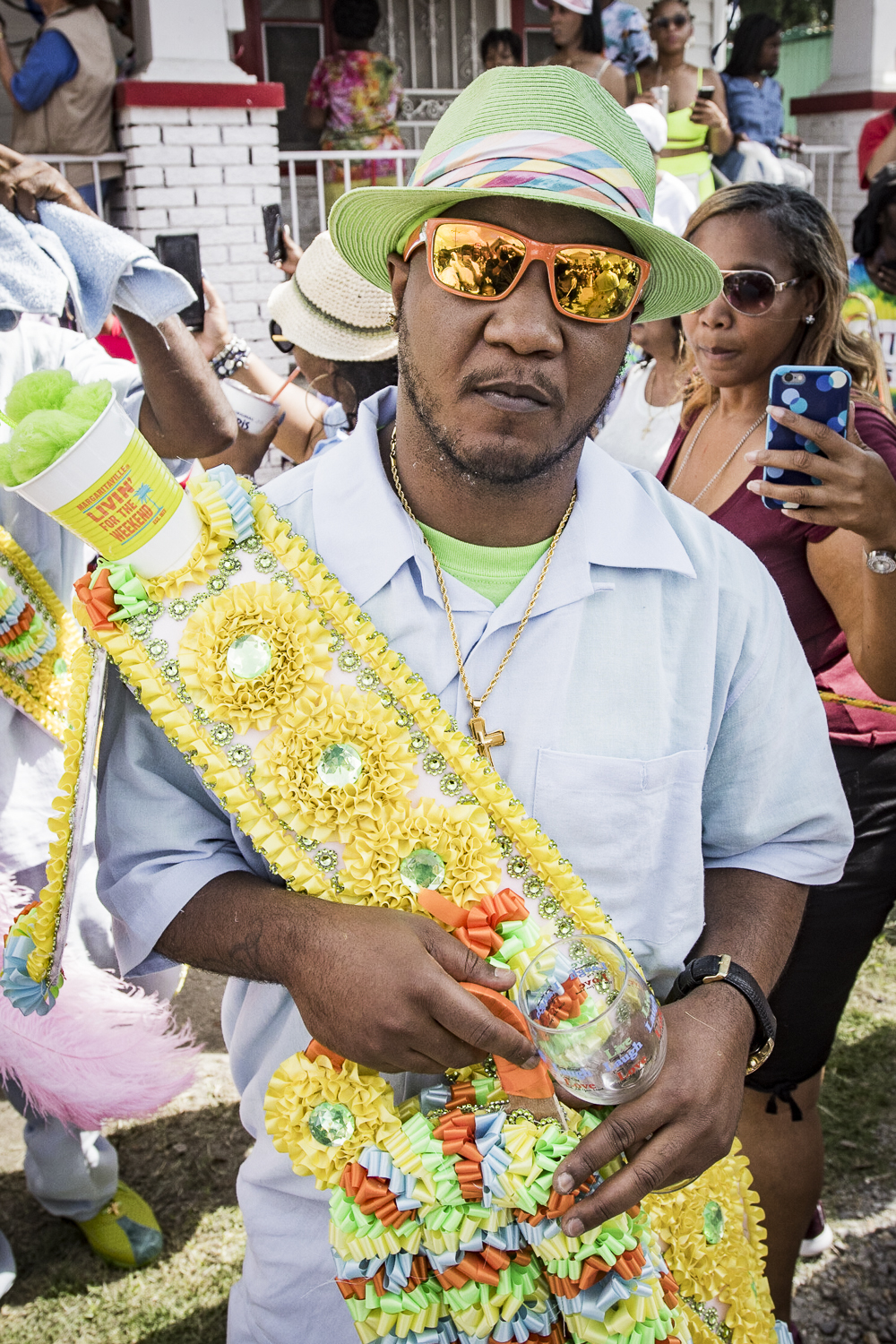 A man in a lime green hat and sunglasses wears a decorated sash and holds a glass during a second line parade.