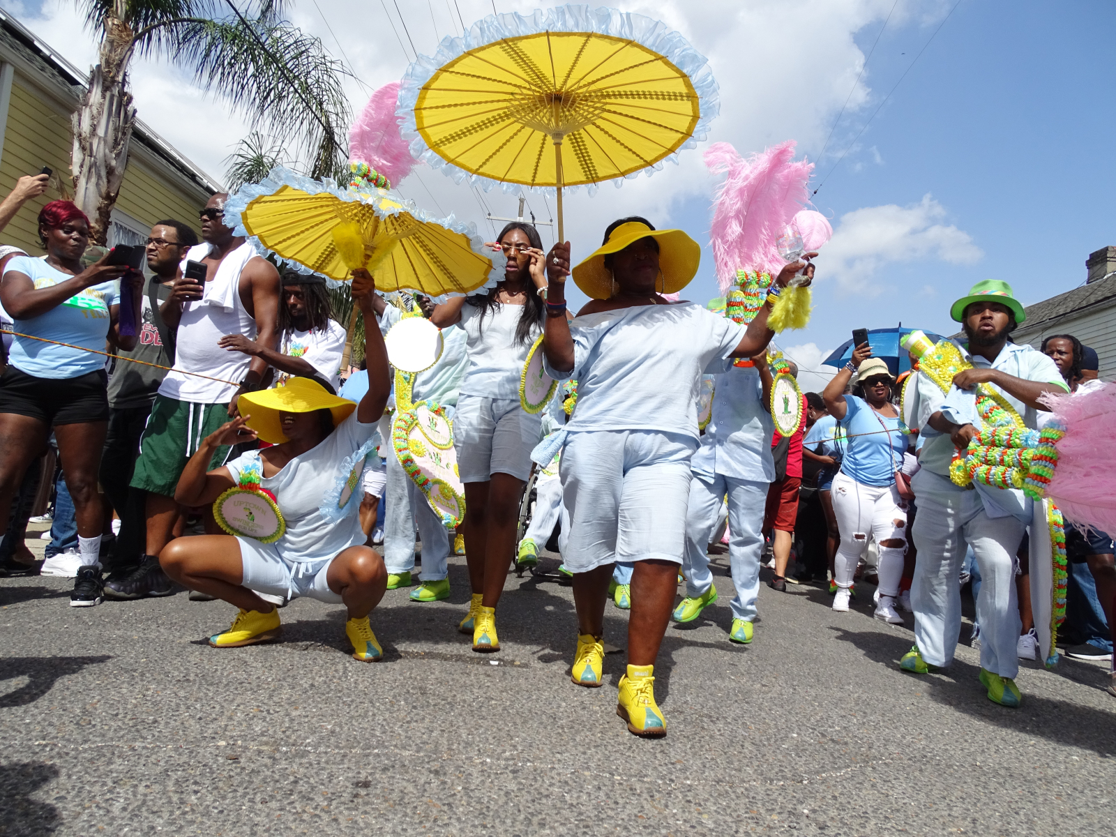 A vibrant second line parade marches down the street, with participants in pastel blue outfits, yellow hats, and matching umbrellas, led by a woman in the foreground.