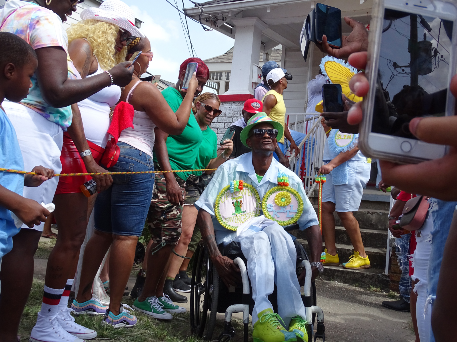 A man in a wheelchair, dressed in a vibrant outfit with decorated medallions, smiles as he moves through a lively second line parade crowd.