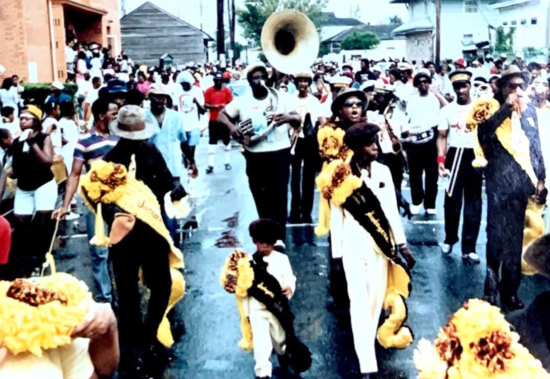 A vintage photograph of a lively second-line parade with elegantly dressed participants wearing black and gold sashes, dancing through the street as a brass band plays behind them. A young child in a matching outfit leads the way, holding a decorated umbrella.