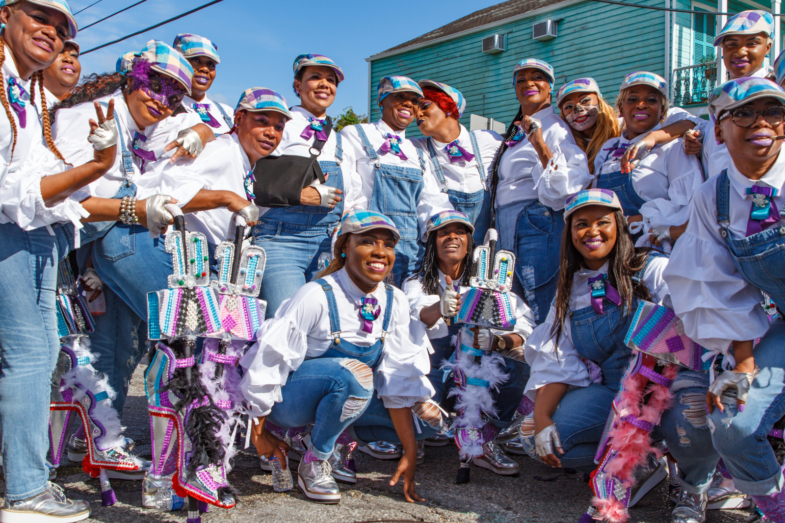 Versatile Ladies of Style pose for a group photo at a second line parade as they celebrate their 10th anniversary. They are wearing matching white shirts, denim coveralls, and denim hats.