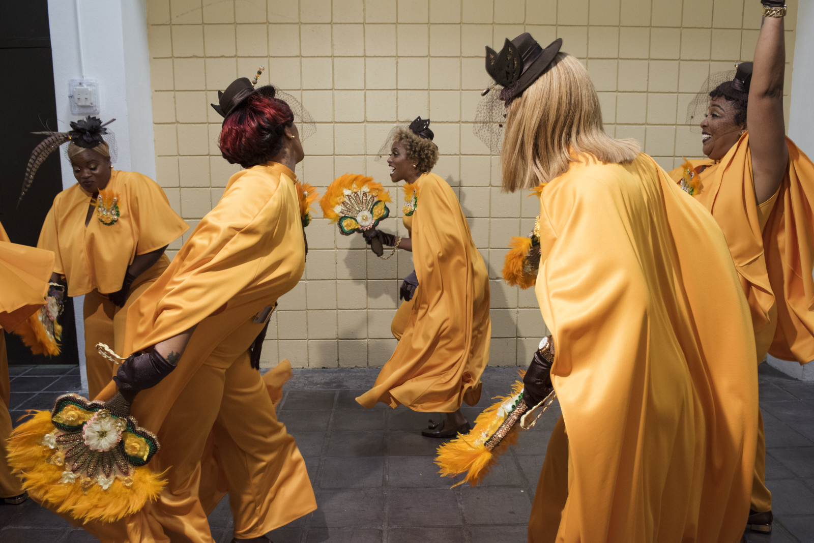 Versatile Ladies of Style dance at the Tremé Center before coming out the door. They are wearing bright orange matching outfits and parade props.