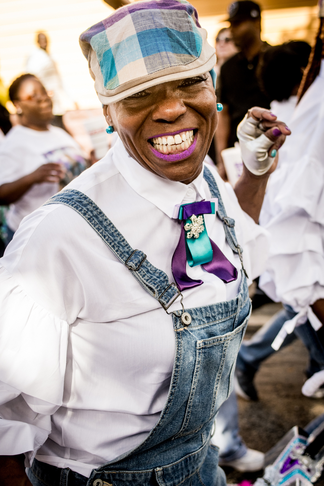 Cheryl Ann Roberts of the Versatile Ladies of Style smiles at the camera at a second line parade. She is wearing a white shirt, denim coveralls, and a denim cap.