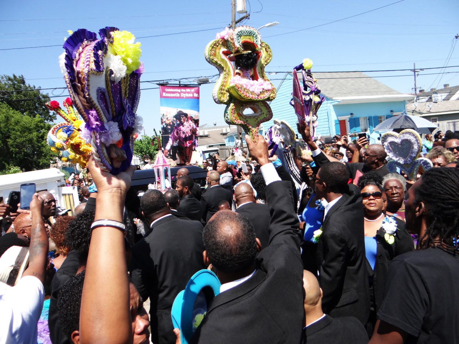 A view from the street as members of the Sudan social aid and pleasure club, wearing suits and raising parade props, honor Kenneth Dykes during his funeral second line parade.