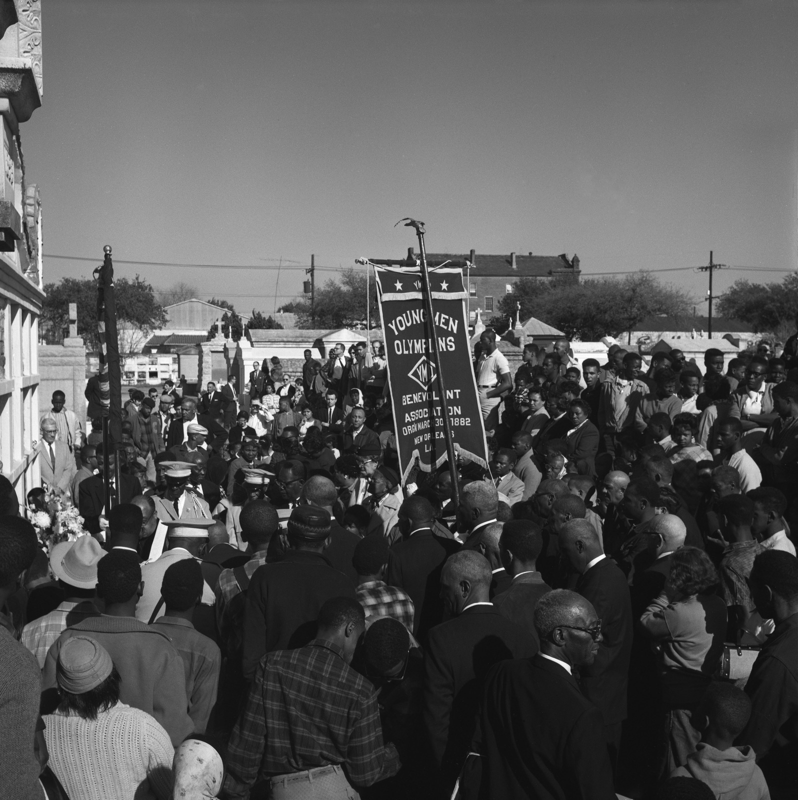 A black and white photo shows the internment of a member at the YMO, Jr. tomb, in Lafayette Cemetery No. 2., with many dressed in suits and hats. A banner reading "Young Men Olympians Benevolent Association, Organized 1882" is held high among the attendees.
