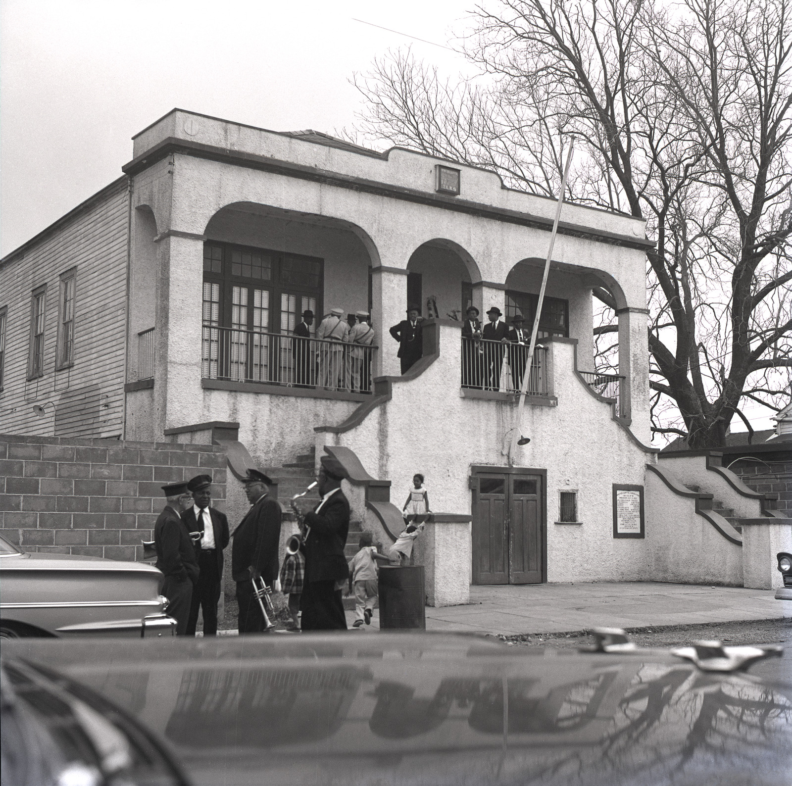 The Eureka Brass Band and YMO, Jr. prepare for a funeral at Elks Hall, 1965. A group of people, dressed formally, gathers on the balcony of a two-story stucco building with arched openings. Below, musicians in dark suits and hats stand near parked cars, one holding a brass instrument.