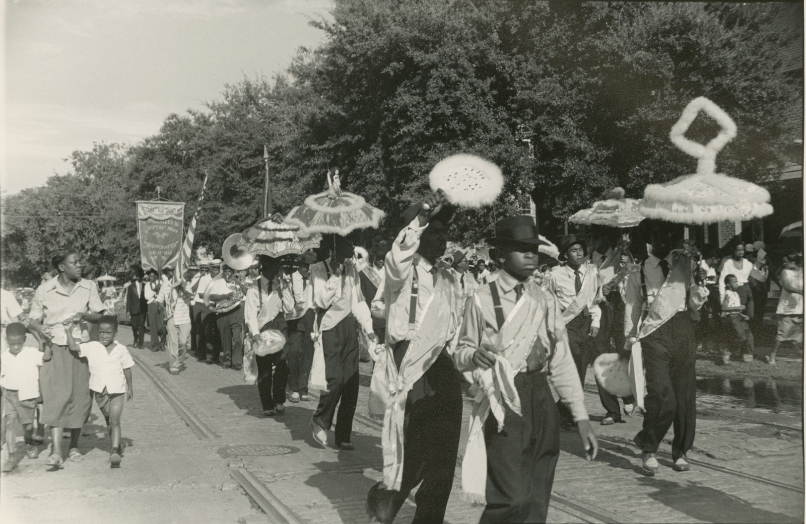 A black and white photo of a YMO, Jr. parade in 1959. A group of young men dressed in formal attire with sashes and hats march in a parade, holding decorated umbrellas. Behind them, a brass band and banner carriers follow, while spectators, including children, walk alongside the procession.
