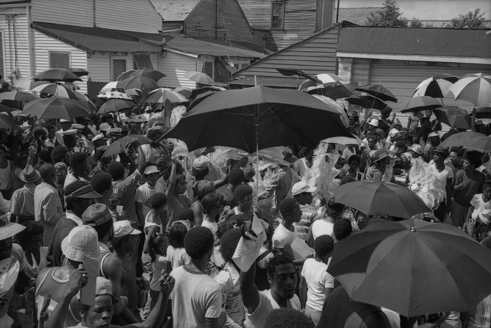 Black and white photo of a YMO, Jr. parade in 1977. A large crowd gathers in the street, dancing and waving umbrellas during a lively second-line parade.